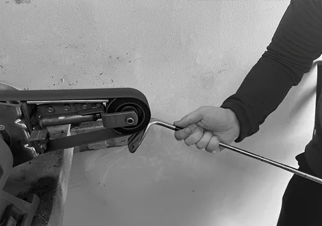 Hands carefully working a golf club shaft on a workshop belt sander